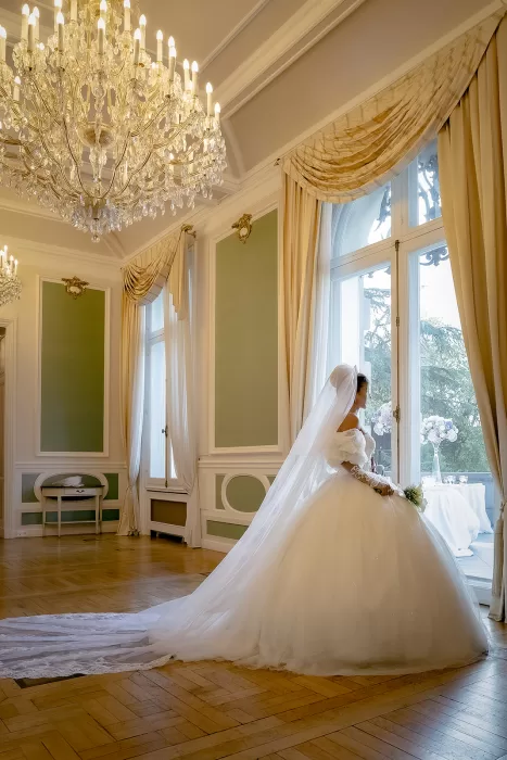 Bride in a voluminous gown standing by tall windows beneath crystal chandeliers inside Château Bouffémont.