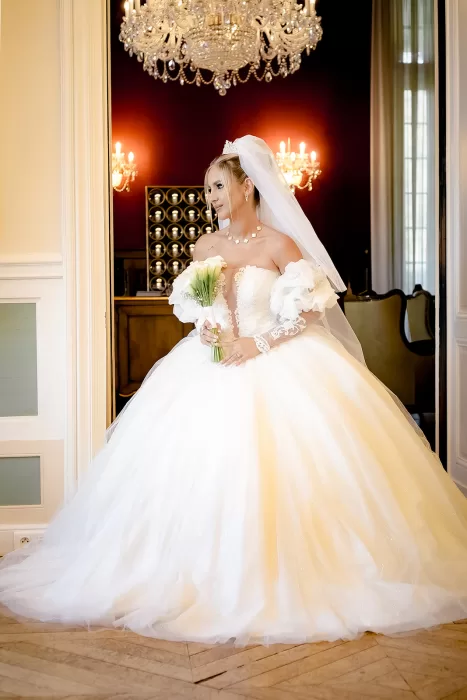 Bride in a voluminous off-shoulder gown standing beneath a crystal chandelier inside Château Bouffémont.