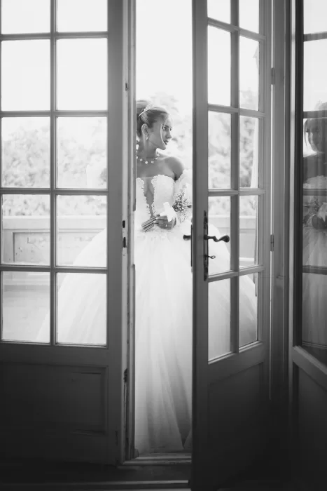 Bride standing behind French doors on the balcony at Château Bouffémont, softly lit in an elegant black and white portrait.