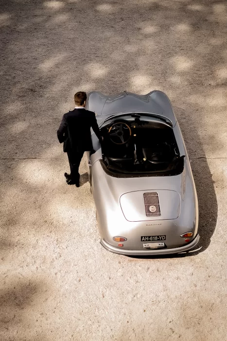Groom in a black tuxedo stepping into a vintage silver Porsche on the gravel driveway at Château Bouffémont.