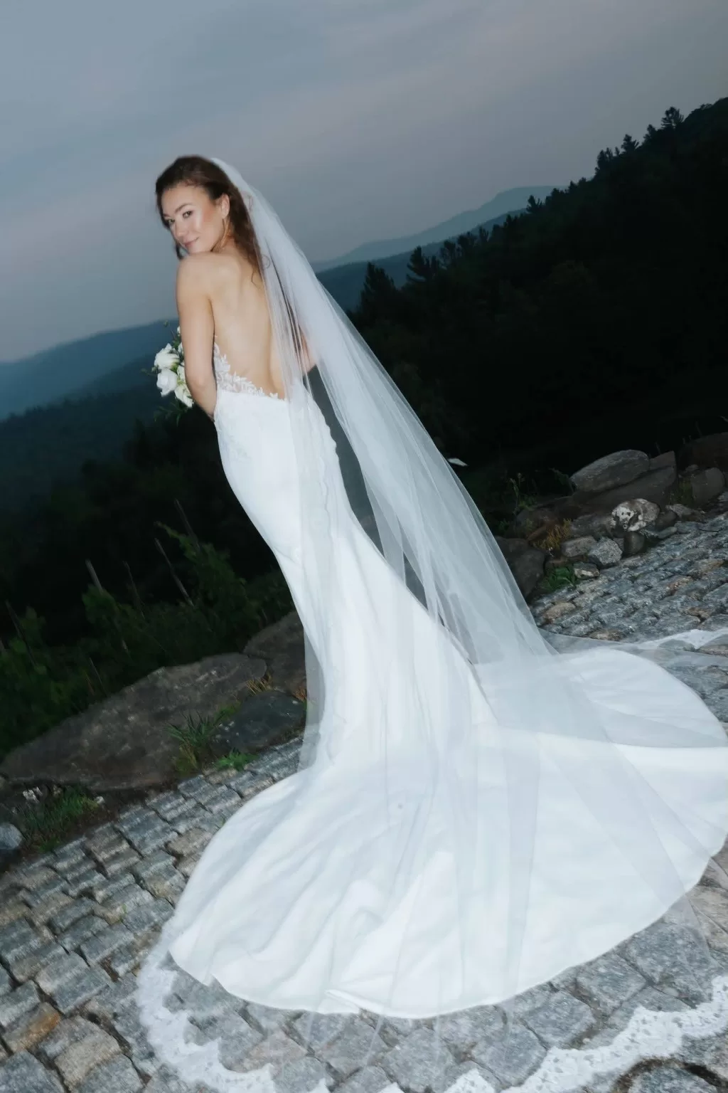Bride looking over her shoulder in a sleek gown and long veil, standing on a stone path with mountain views at Château Ste. Agnès.