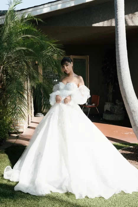 Bride standing in the garden outside Common Goods wearing a voluminous off-the-shoulder wedding gown surrounded by palm trees.