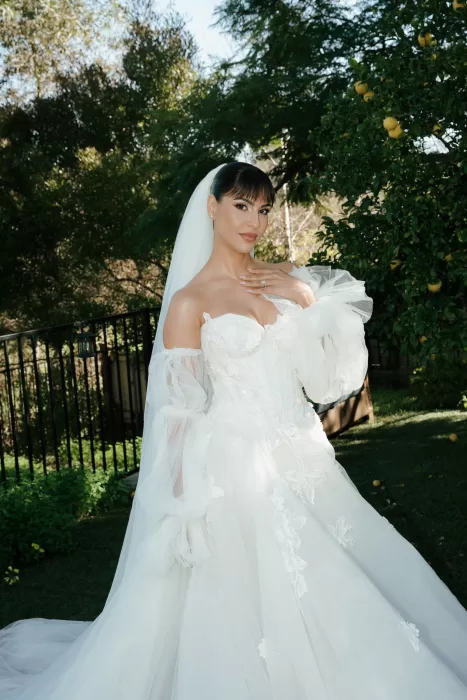 Bride standing in the garden outside Common Goods wearing a voluminous off-the-shoulder wedding gown surrounded by palm trees.