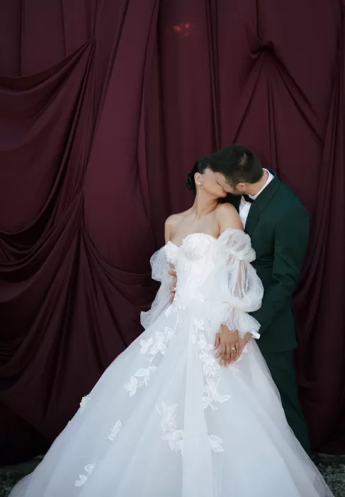 Bride and groom embracing and kissing in front of burgundy drapery backdrop at Common Goods.