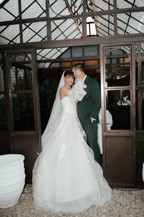 Bride and groom embracing inside the glass greenhouse at Common Goods, framed by iron doors and warm string lights.