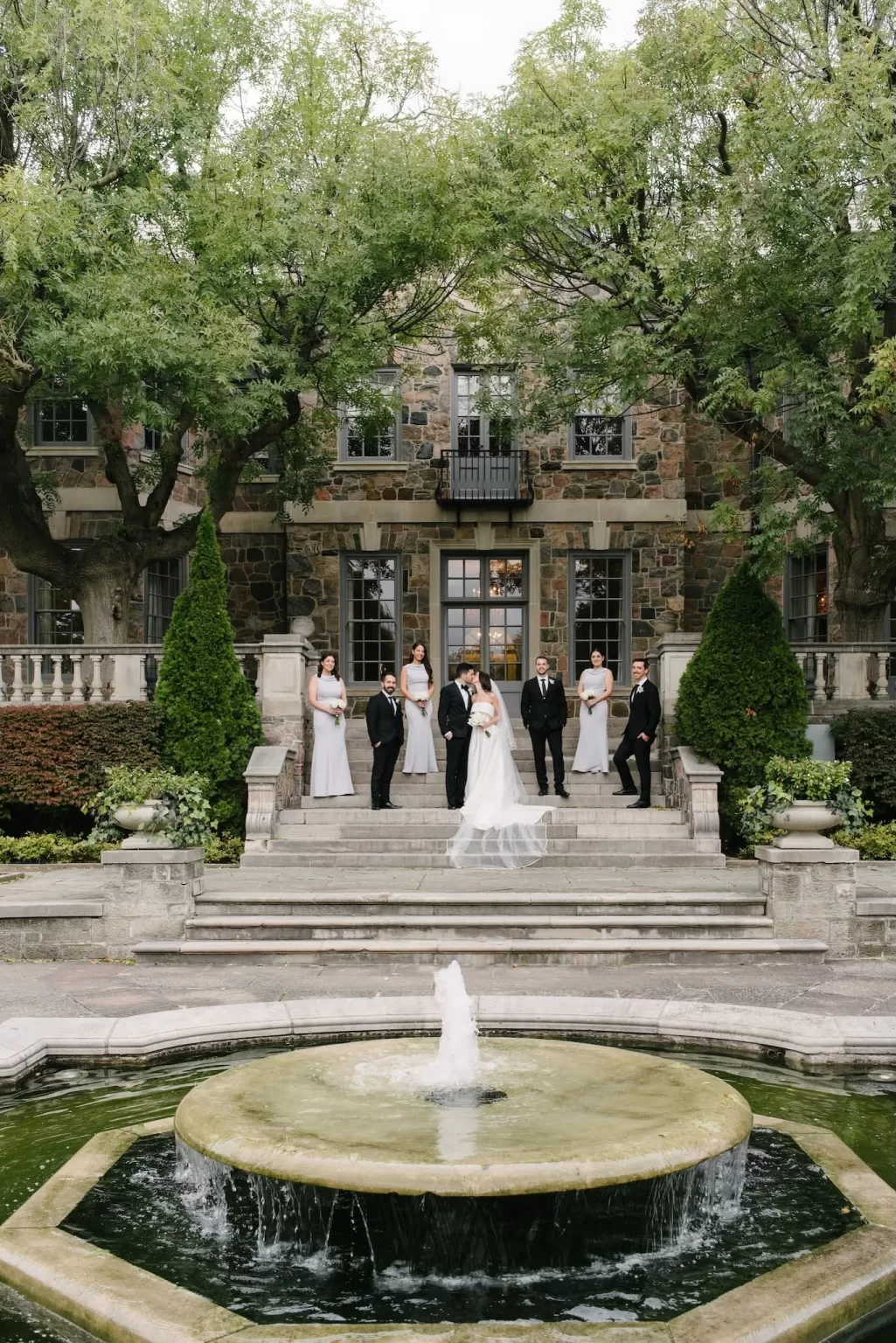 Bride and groom with their wedding party gathered by the fountain and stone steps of Graydon Hall.