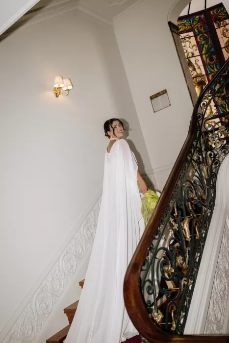 Bride standing on the ornate staircase at Infante Sagres holding a green floral bouquet, her long cape flowing behind her.