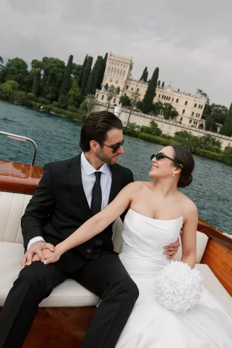 Bride and groom sitting on wooden boat with Isola del Garda villa in the background on Lake Garda