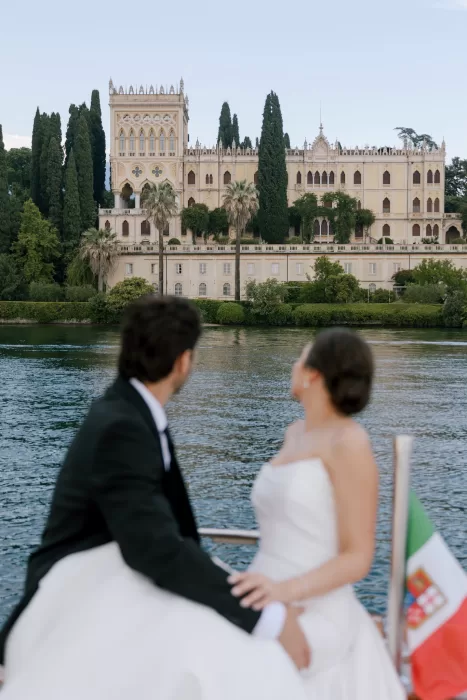 Bride and groom on boat looking toward Isola del Garda villa across Lake Garda