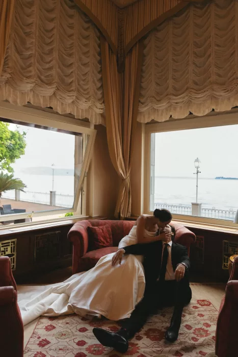 Bride leaning over groom for kiss on red sofa with lake views through windows at Isola del Garda