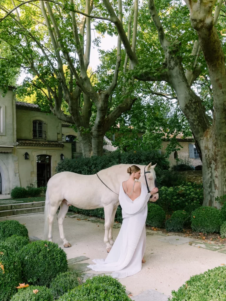 Bride in sculptural gown standing beside a white horse beneath plane trees in the gardens of Mas de Chabran.