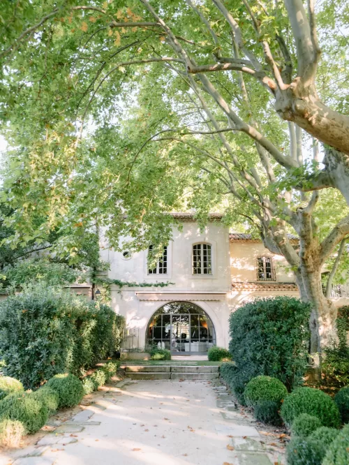 Sunlit facade of Mas de Chabran framed by tall plane trees and manicured garden paths.