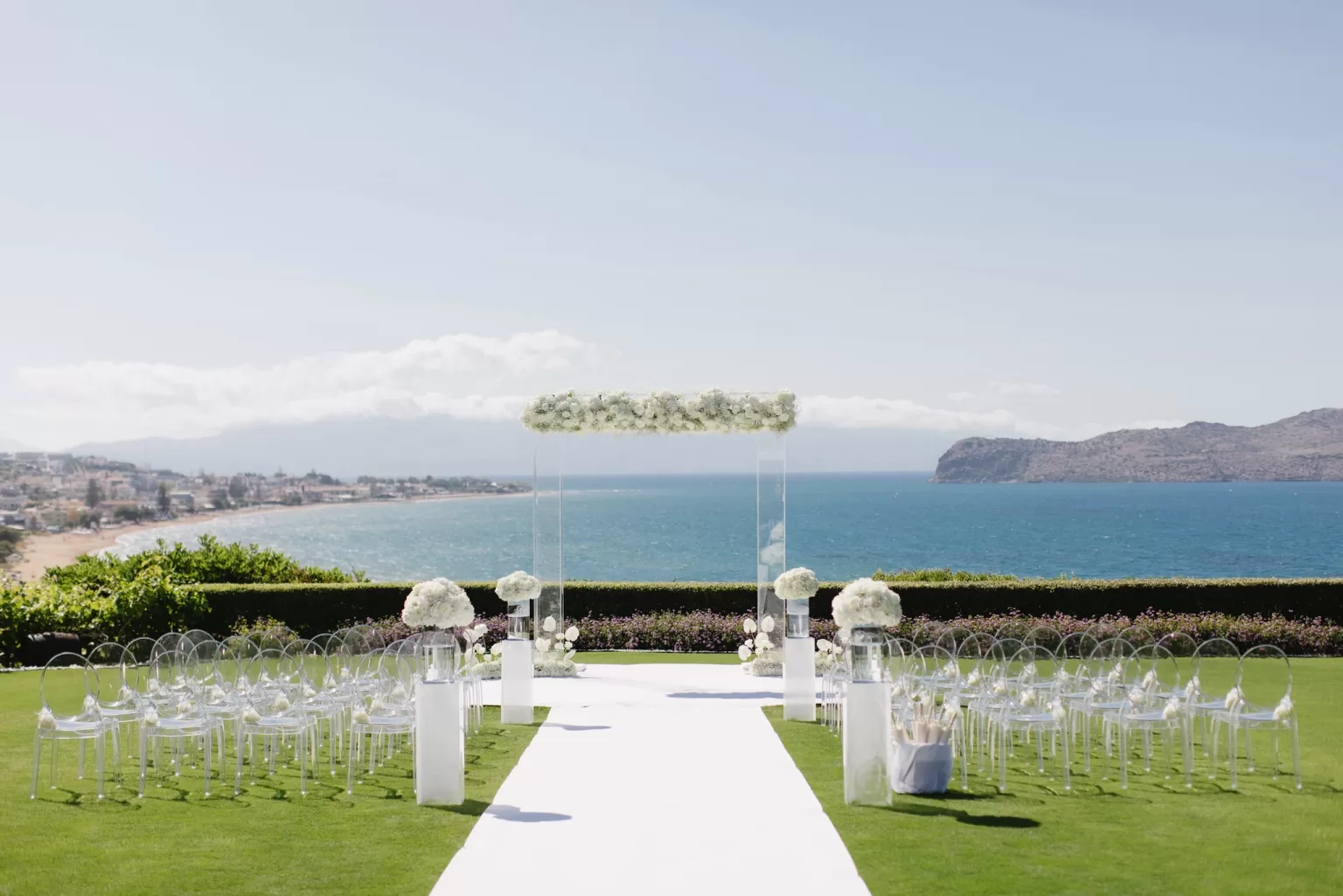 Modern glass arch ceremony at Sea View by En Kipo with clear chairs and white floral aisle against a coastal backdrop