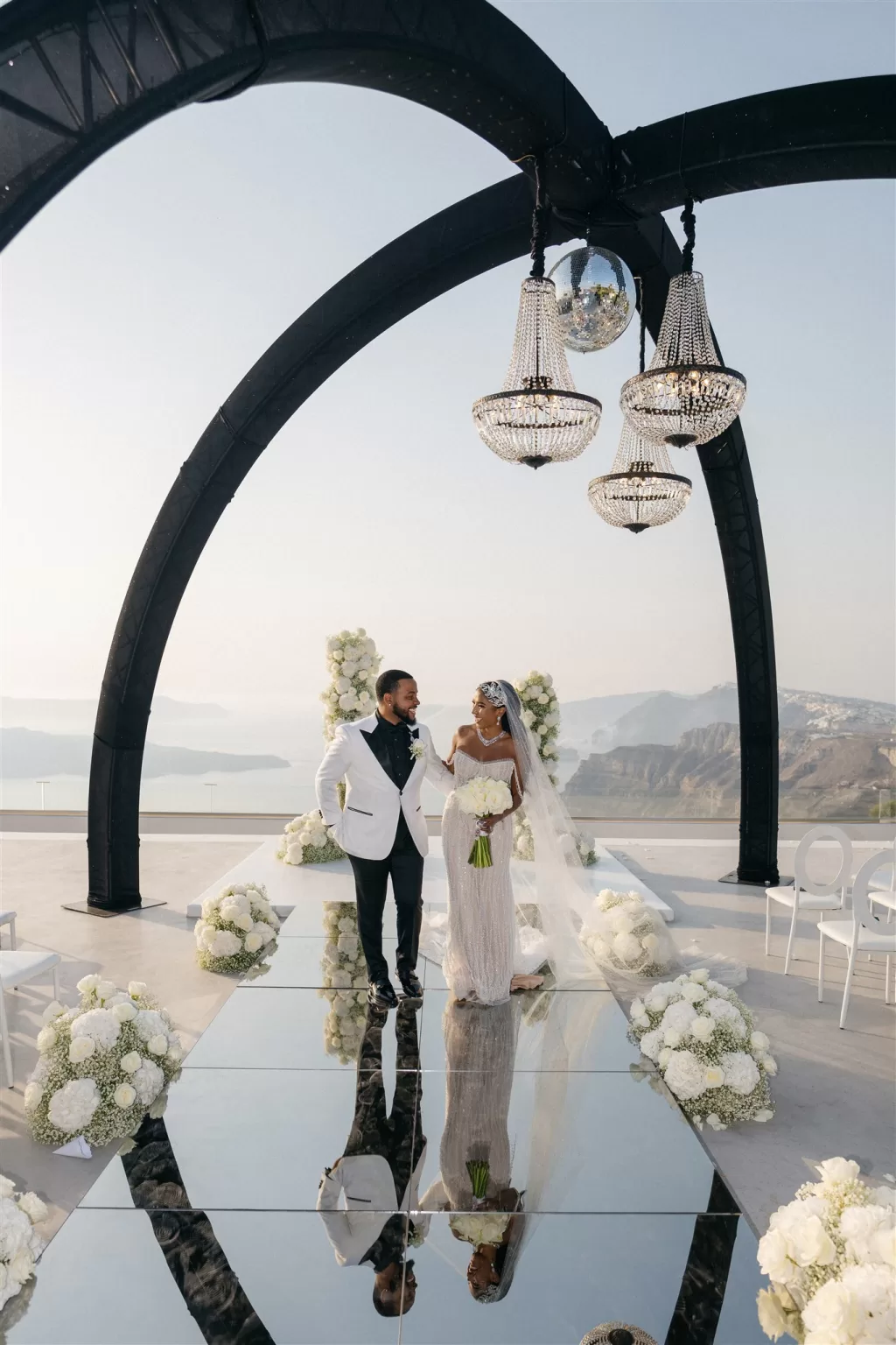 Bride and groom walking together under black ceremony arch with floral arrangements and sea views