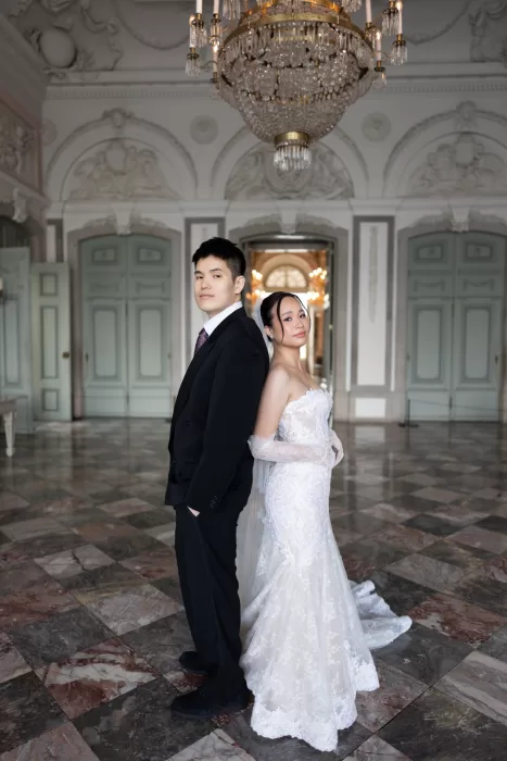 Bride and groom standing back to back beneath a grand chandelier in an elegant historic hall
