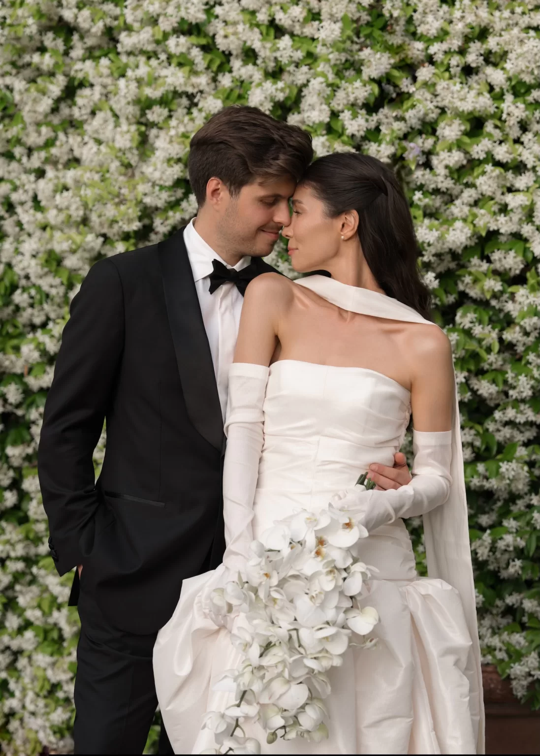 Bride and groom embracing in front of a flowering wall, with the bride holding a cascading white orchid bouquet.