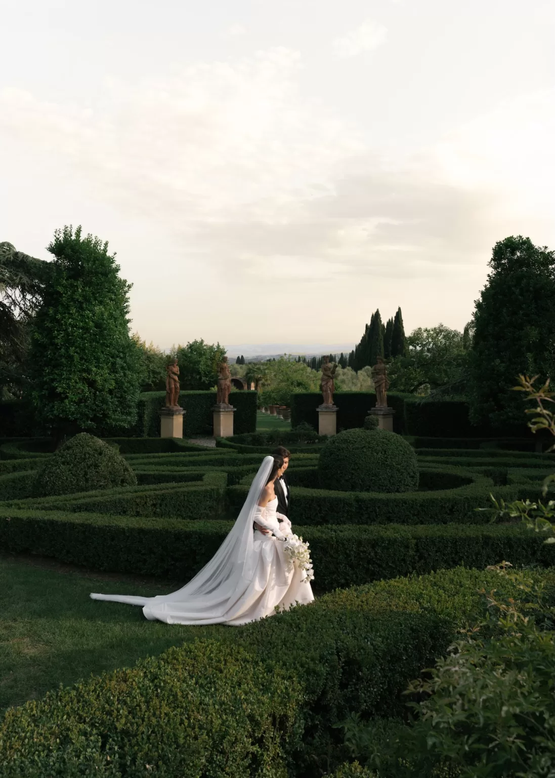 Bride and groom strolling through manicured Italian garden with sculpted hedges at sunset