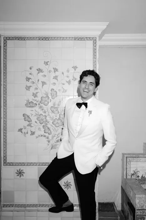 Groom in white tuxedo standing by decorative tiled wall at Finca Morneta