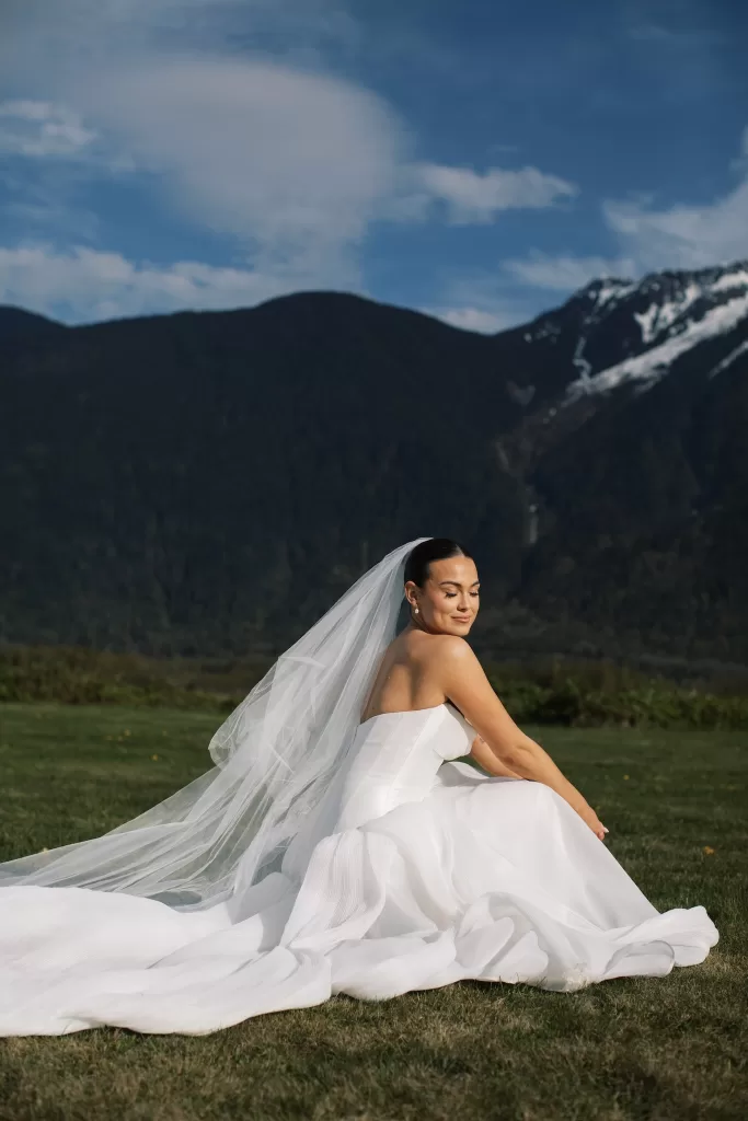 Bride seated on grass with veil and mountain backdrop at Fraser River Lodge