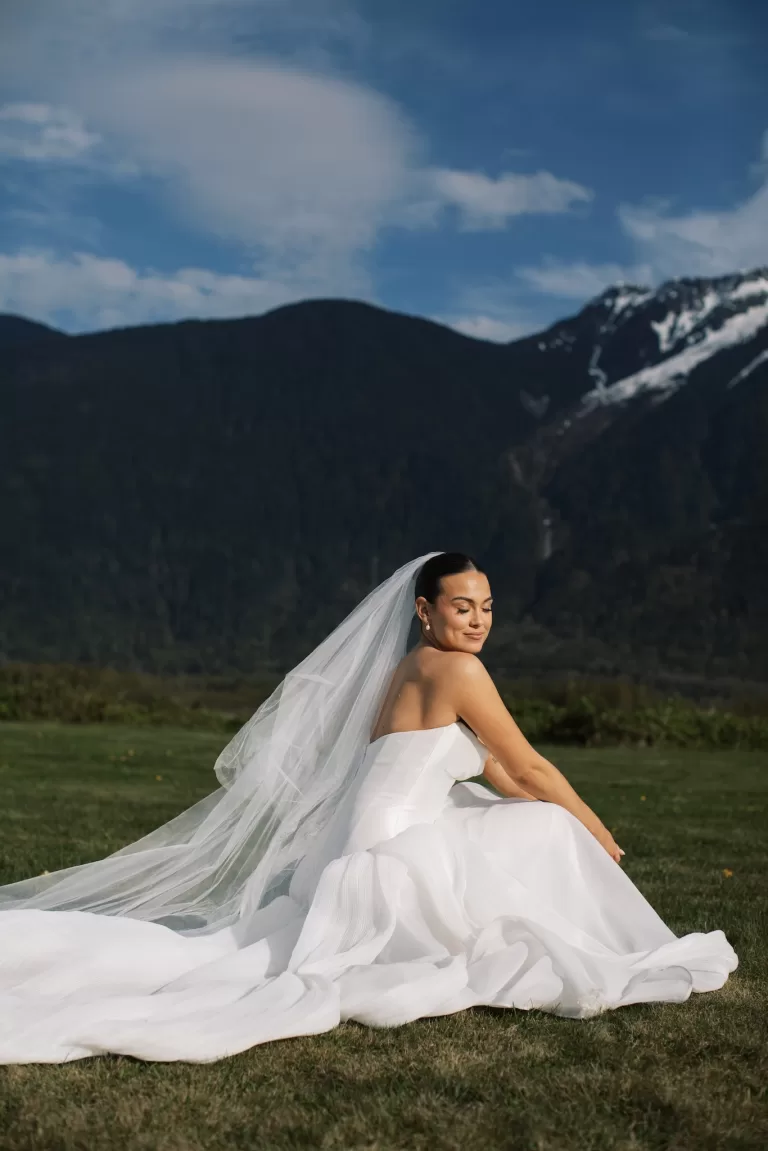 Bride seated on grass with veil and mountain backdrop at Fraser River Lodge