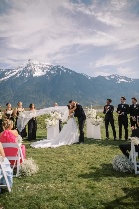 Bride and groom sharing first kiss during outdoor ceremony with mountains behind at Fraser River Lodge