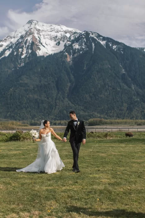 Bride and groom walking hand in hand across lawn with mountain backdrop at Fraser River Lodge