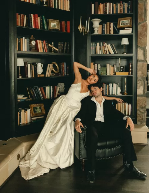 Relaxed editorial portrait of the couple beside built-in bookshelves and a leather chair.