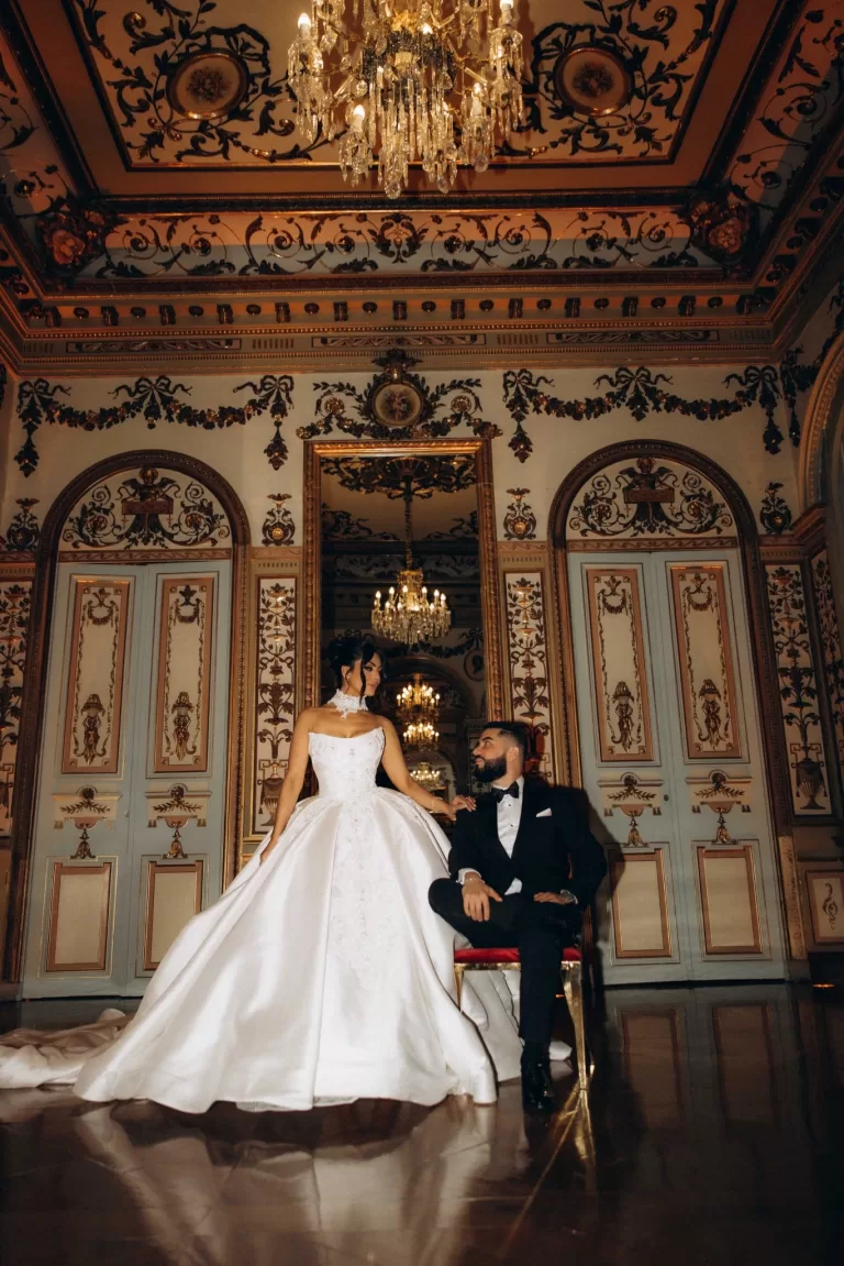 Bride standing beside the groom seated on a velvet chair in the ornate grand salon of Palacio Metropolitano beneath crystal chandeliers.