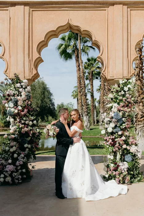 Bride and groom embracing beneath floral ceremony arch with palm trees at Palais Namaskar