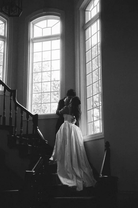 The couple kiss on the staircase, framed by grand windows and soft shadow.