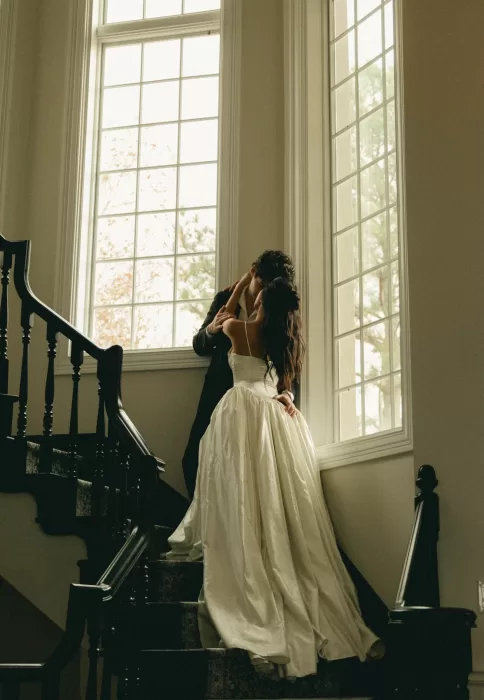The couple kiss on a staircase beneath tall windows and soft afternoon light.