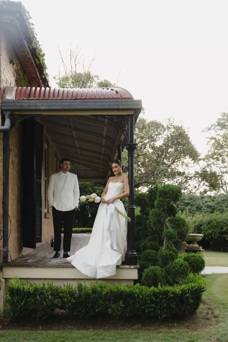Bride holding lotus bouquet while standing on the veranda with groom at Wallalong House.