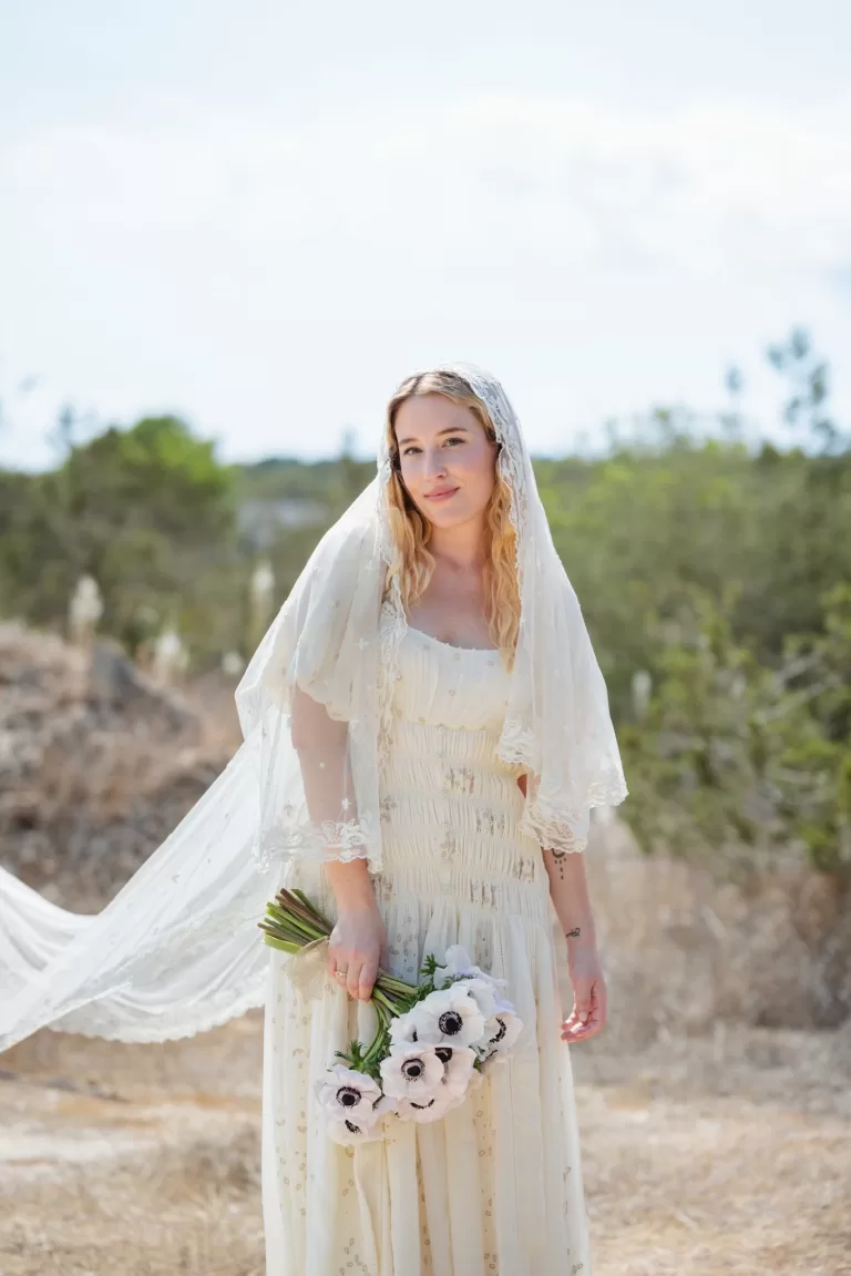Bride in a soft ivory gown and lace-edged veil holding a white anemone bouquet in the Ibiza countryside.