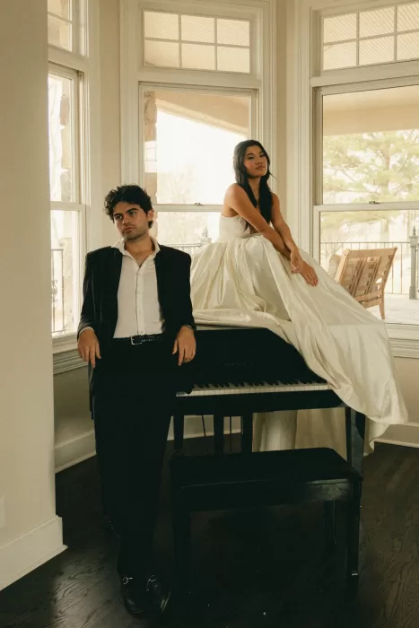 Editorial portrait of the couple by tall windows and a glossy black piano.