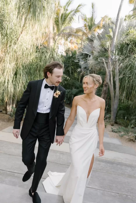 Bride and groom walking hand in hand through lush palms in Baja.