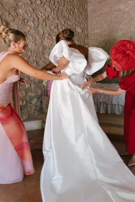 Bride being helped into gown by family, intimate preparation moment in warm interior