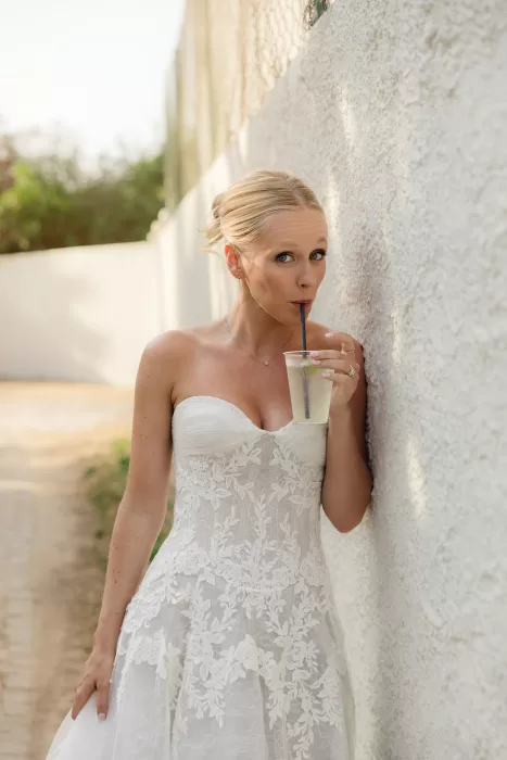 Bride sipping a cocktail beside a whitewashed wall in warm afternoon light.