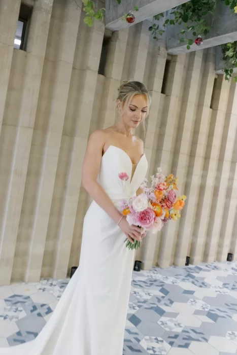 Bride holding a pink and orange bouquet against a pale ribbed architectural wall.