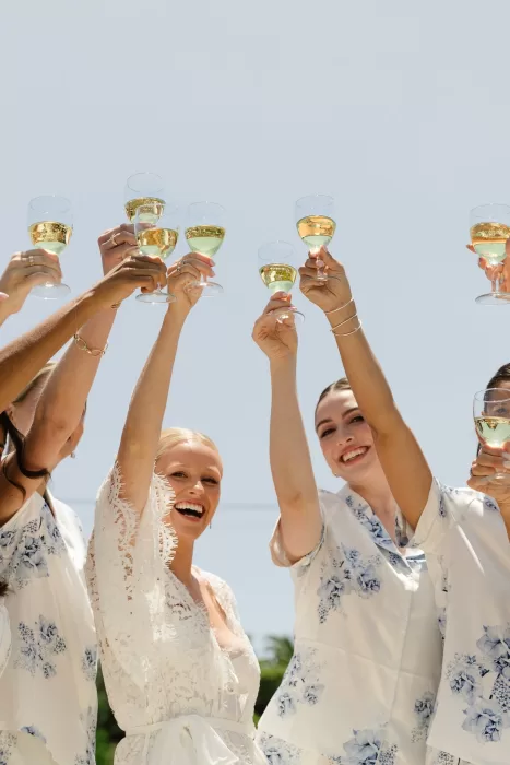 Bride and bridesmaids raising champagne glasses against a clear coastal sky.