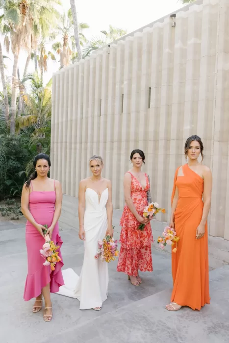 Bride and bridesmaids holding colourful bouquets against a pale architectural wall.