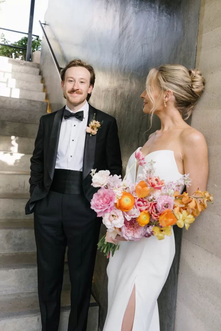 Groom smiling at the bride as she holds colourful flowers by the staircase.