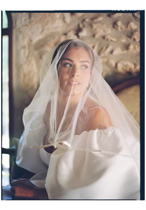 Soft bridal portrait by window, veil draped gently, warm light against textured stone walls