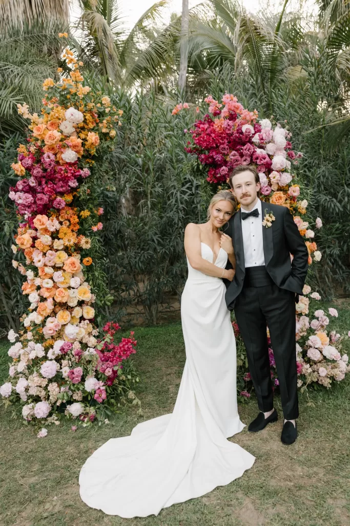 Bride and groom standing together between vivid floral arches in a tropical garden.