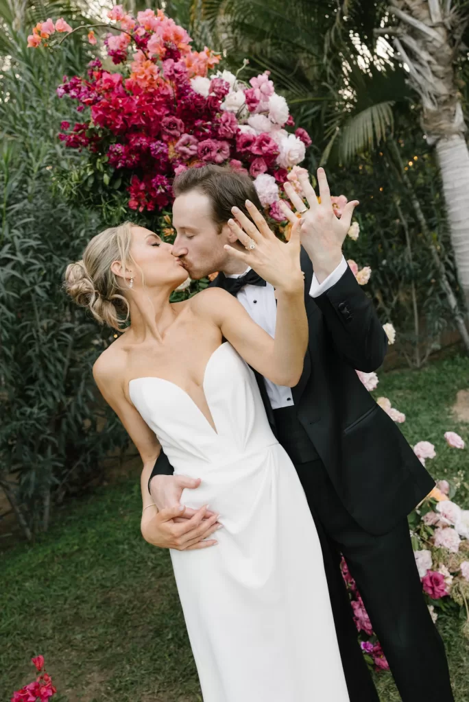 Newlyweds kissing in front of a vivid floral installation and tropical garden greenery.
