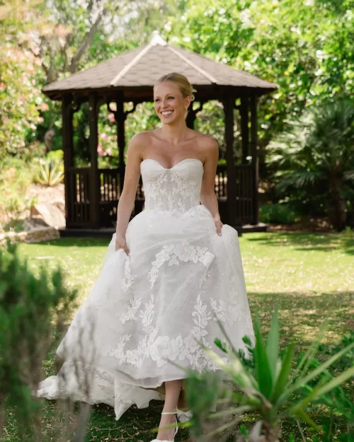 Bride walking through a shaded garden in a floral lace wedding gown.