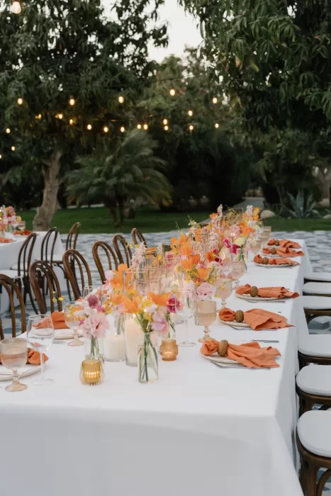 Long dinner table with colourful flowers, terracotta napkins, and glowing candles outdoors.