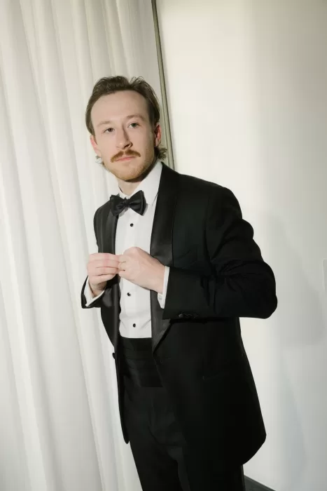 Groom in a black tuxedo adjusting his jacket in a softly lit room.
