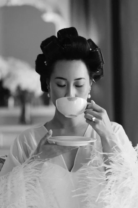 Bride in feathered robe sipping tea during a quiet black-and-white getting-ready moment.