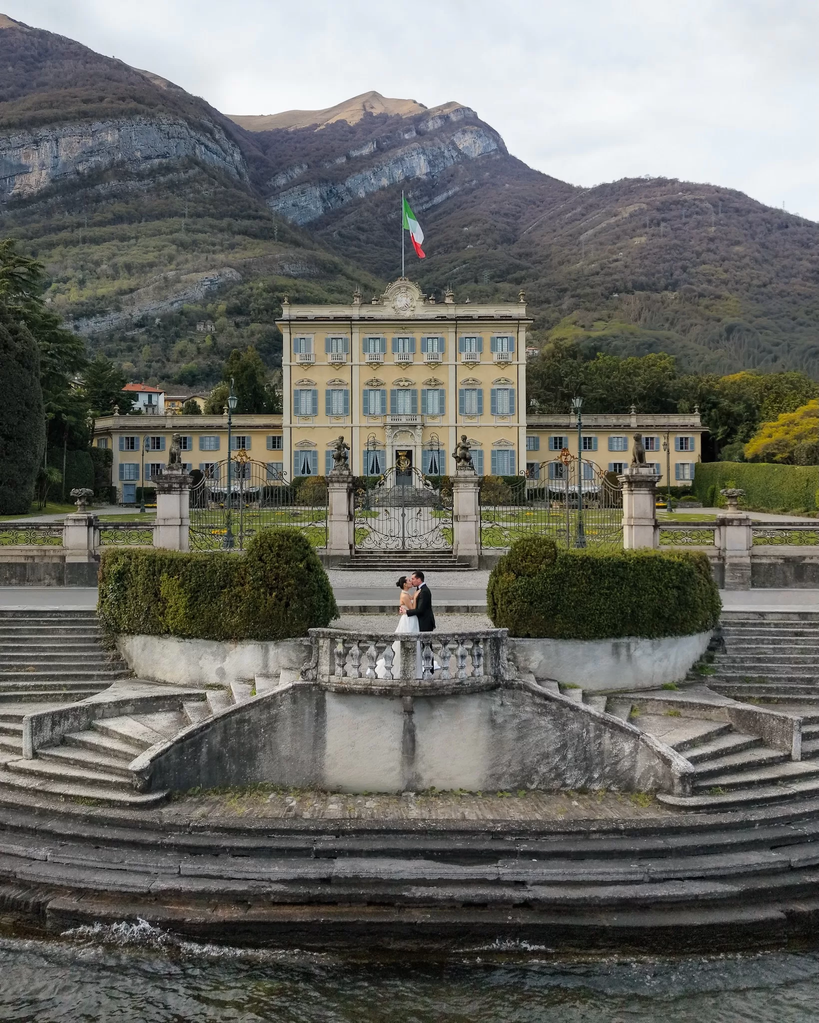 Couple kissing on the lakefront terrace, framed by Hotel Villa Marie and steep mountains.
