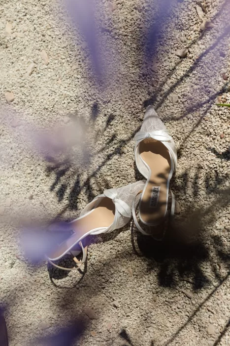 Delicate lace bridal shoes arranged on gravel beneath soft lavender shadows.